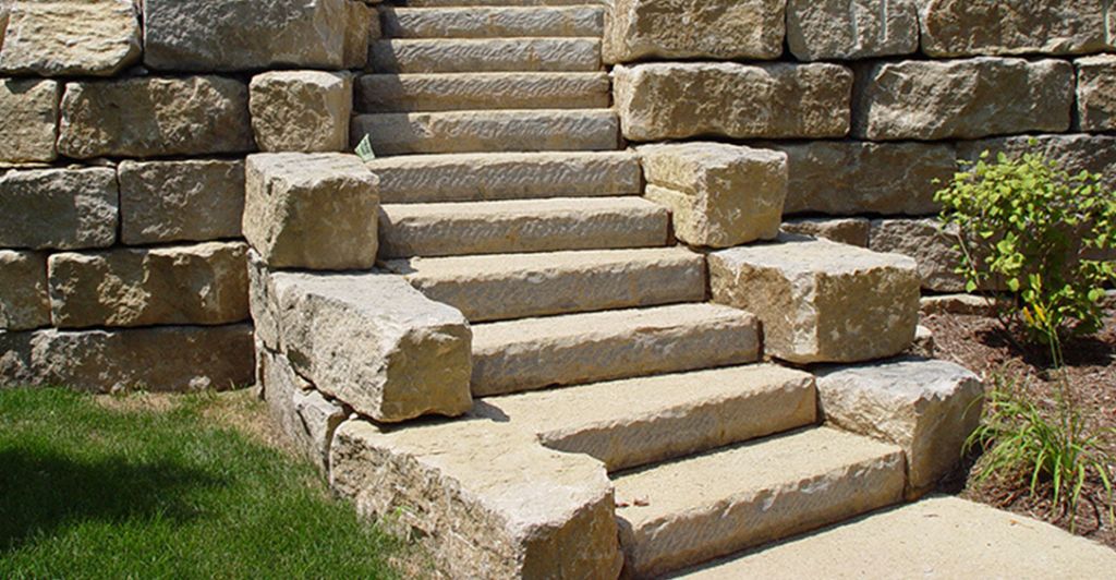 Limestone steppers, set alongside a large outcropping wall, create a natural and rustic pathway, seamlessly blending with the surrounding landscape for an elegant yet functional outdoor design in Des Moines
