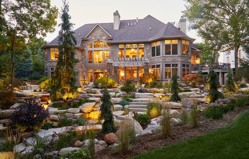 A beautiful backyard landscape design with flagstone pathways creates a natural and inviting atmosphere. Large boulder wall fountains and a meandering stream add soothing water elements, while strategically placed evergreen trees provide year-round structure and greenery. Surrounding the space, perennial plantings offer vibrant seasonal blooms and texture, enhancing the overall aesthetic with pops of color and movement. This design creates a tranquil outdoor retreat that blends natural materials with lush greenery, perfect for relaxing or entertaining by Ted Lare Design Build in Des Moines