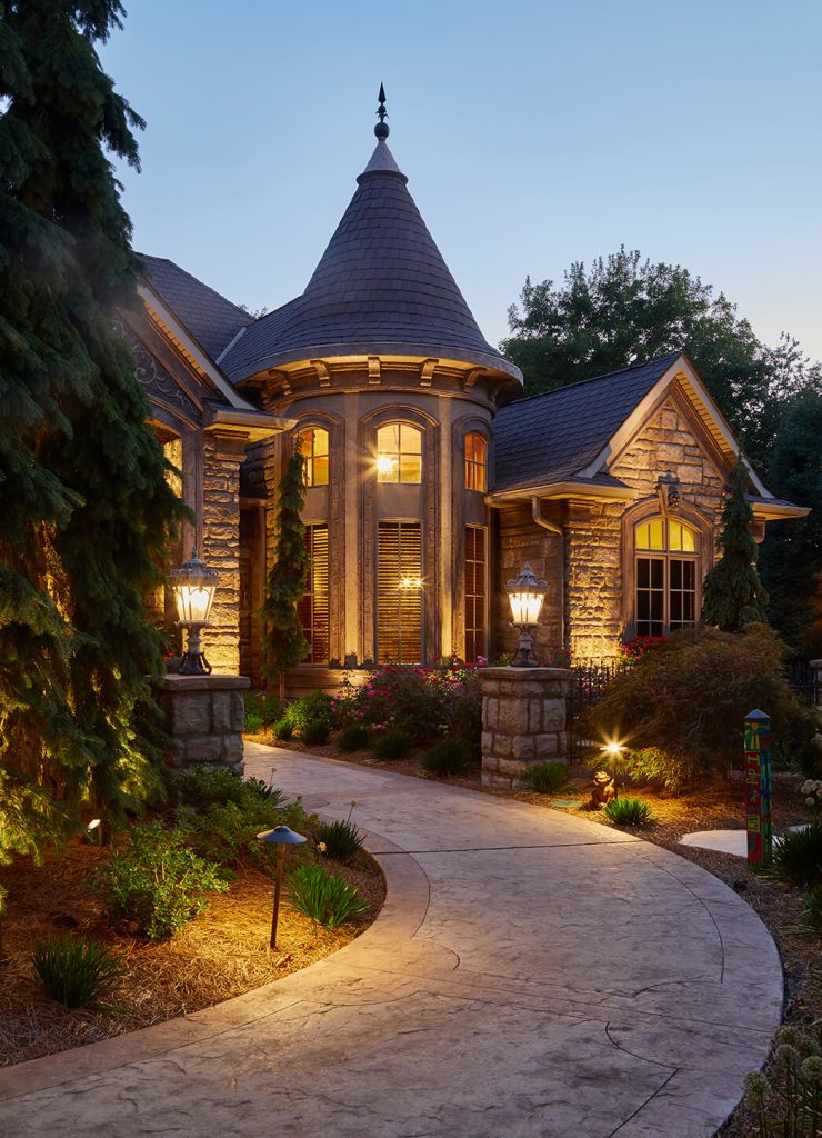 A stunning front yard landscape design featuring concrete pathways leads visitors through the space, accented by Unilock stone edging that provides a clean and elegant border. Large evergreen trees create a strong visual presence, offering year-round greenery and structure. Surrounding these trees, a variety of perennial plantings bloom throughout the seasons, adding pops of color and texture. This design combines functionality and beauty, making the front yard a welcoming and visually striking space.