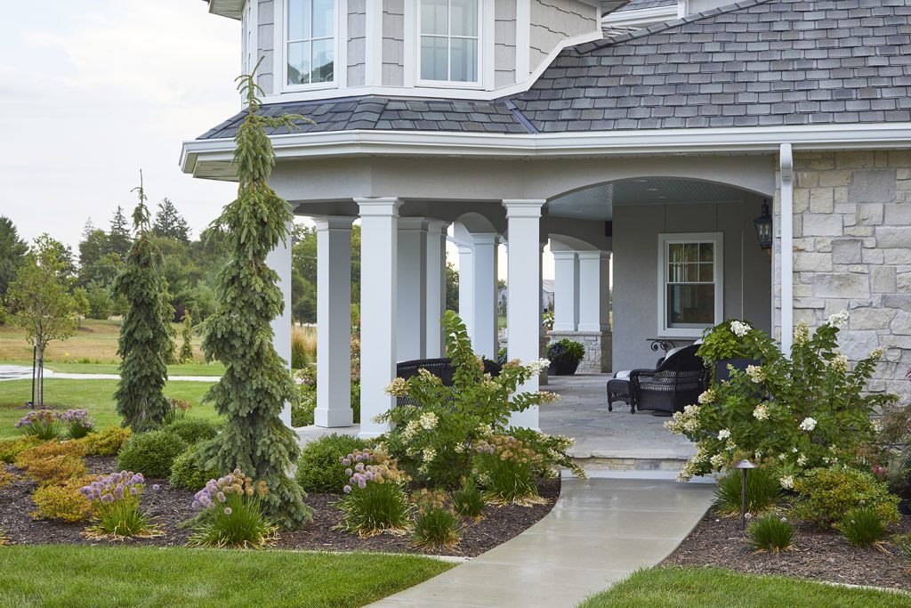 Outdoor landscaping by Ted Lare Design Build showcases Unilock stone edging that frames the space beautifully, complemented by stately evergreen trees for year-round structure. Perennial plantings provide vibrant seasonal color, while a sleek cube accent adds a modern touch. A thoughtfully crafted summer planter design completes the scene, enhancing the landscape with dynamic layers of texture and blooms.