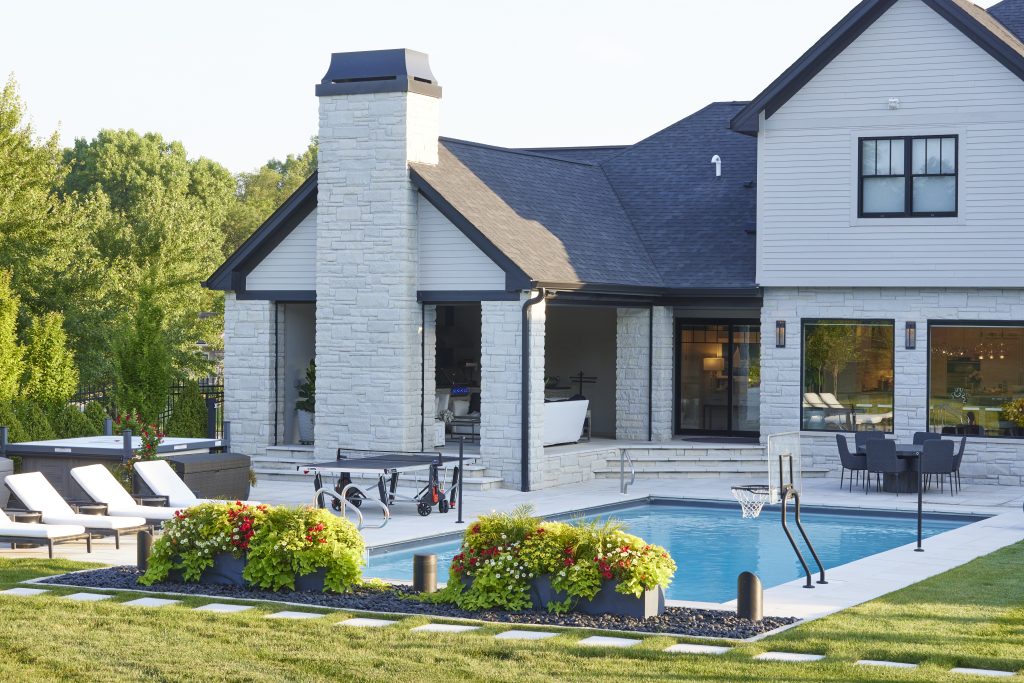 Unilock stone pool deck and patio with stone walls and steppers creates a timeless and functional outdoor space, offering both elegance and durability. The stone wall adds structure and a sense of privacy, while the steppers provide a natural, path-like flow across the area, connecting different spaces. The colorful planter pot design brings a pop of vibrancy with blooming flowers, adding seasonal color and texture. Incorporating beach pebbles and additional steppers in sod creates a relaxed, beach-inspired atmosphere that complements the pool area, making it an ideal spot for lounging and enjoying the outdoors by Ted Lare Design Build