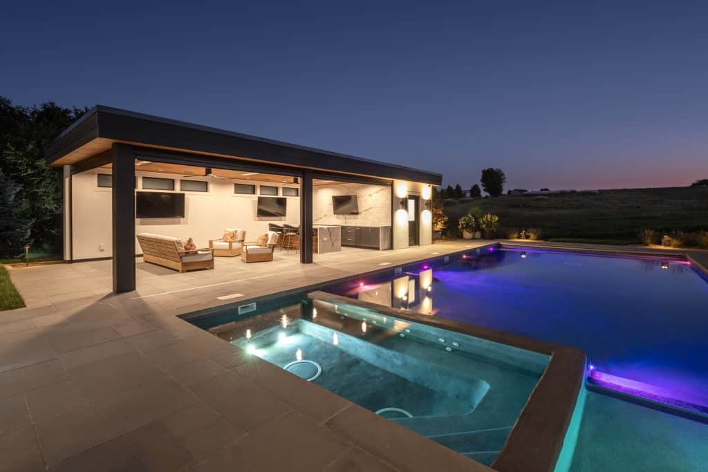 An outdoor pool patio designed with Unilock pavers and custom stonework, complemented by a beautiful pool house featuring an outdoor kitchen, makes for a luxurious and functional backyard space. The design by Ted Lare Design Build incorporates high-quality materials like natural stone and durable pavers to create a cohesive, stylish outdoor environment. The pool area is surrounded by lush landscape plantings, including vibrant perennial flowers, shrubs, and trees, adding color and texture throughout the seasons in Des Moines with landscape lighting